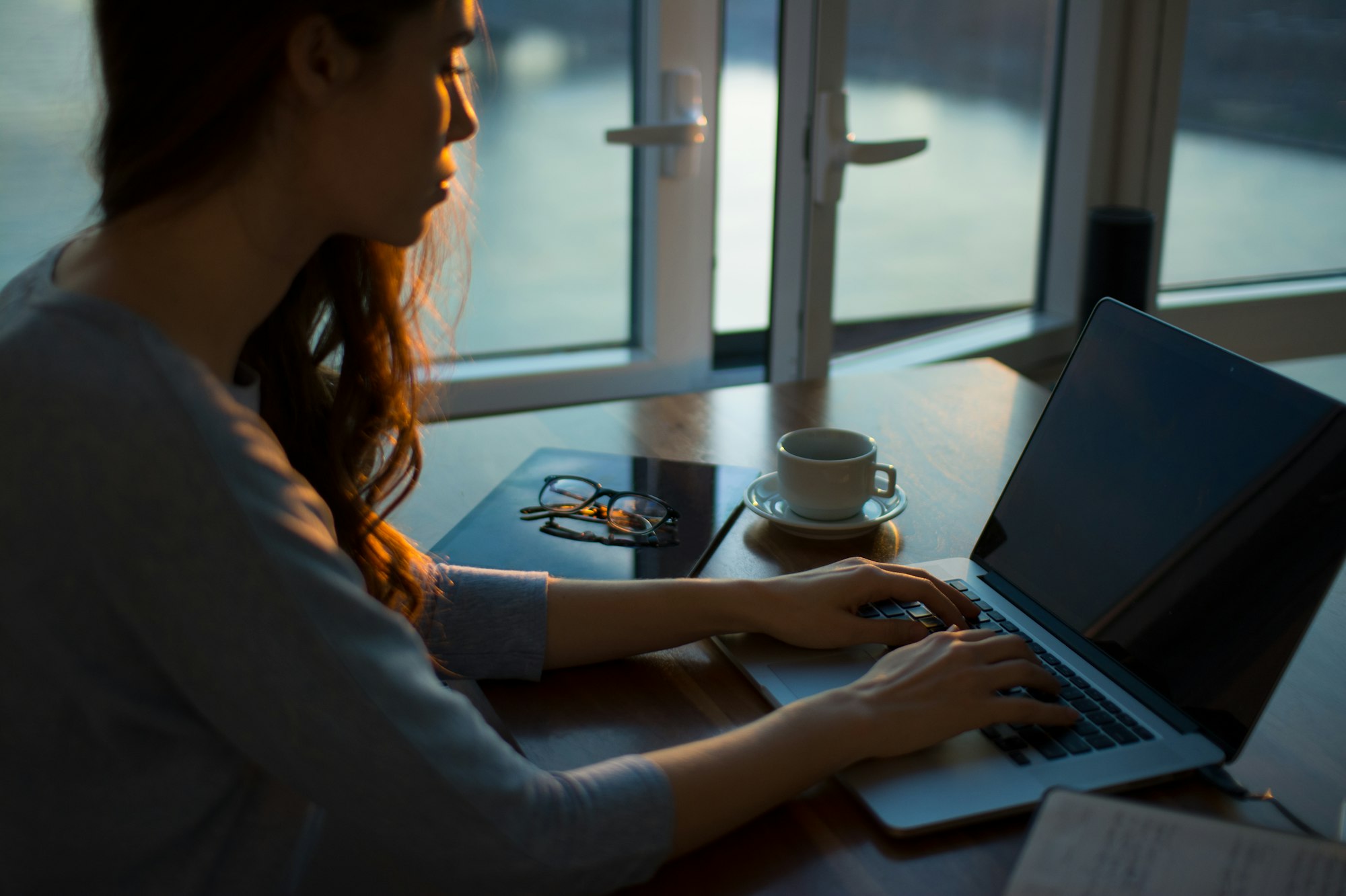 A person working on a laptop at a table beside a window in a dim office
